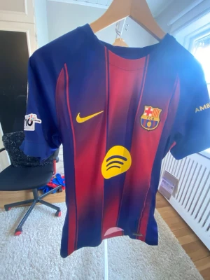 Barcelona matchtröja  - Säljer en officiell FC Barcelona matchtröja i blått och rött med gula detaljer. Tröjan har klubbmärke, Nike-logga och sponsortryck på bröstet samt tryck på ärmar och rygg. Tillverkad i lätt och ventilerande material, perfekt för fotbollsträning eller match. Köpt på Barca shopen i Spanien 