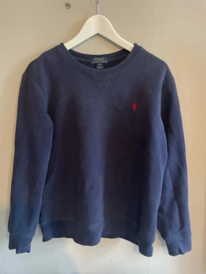 Mörkblå sweatshirt från Polo Ralph Lauren - Klassisk mörkblå sweatshirt från Polo Ralph Lauren med rund halsringning och den ikoniska röda logotypen broderad på bröstet. Tröjan har långa ärmar och ribbade muddar vid ärmslut och nederkant. Perfekt för en avslappnad och stilren look.