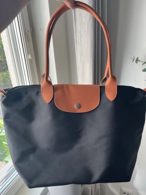 Svart tote handväska med bruna detaljer - Stilren svart tote handväska med bruna handtag och lock i läderimitation. Väskan har en klassisk form, dragkedja upptill och knappstängning på locket. Perfekt storlek för att bära med sig det viktigaste och passar till många olika outfits. Den är köpt på en marknad i Grekland för ungefär 65 euro. Använd bara en gång.