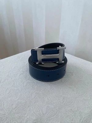 Mörkblått skärp med silverdetalj - Snyggt mörkblått skärp med stor H-formad silverspänne och vita kontrastsömmar. Skärpet har en stilren design och passar perfekt till jeans eller kostymbyxor. Tillverkat i skinn för en exklusiv känsla.