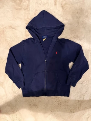 Mörkblå hoodie från Polo Ralph Lauren - Mörkblå hoodie med dragkedja från Polo Ralph Lauren. Klassisk design med huva, två fickor framtill och den ikoniska röda logotypen broderad på bröstet. Tillverkad i mjukt bomullsmaterial som är skönt att bära. Perfekt för en avslappnad och stilren look.