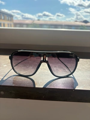 Svarta pilotbågar solglasögon Carrera - Snygga solglasögon från Carrera med klassisk pilotform och svarta bågar. Glasen är tonade i en mörk nyans som ger en cool look. Perfekta för soliga dagar och ger en sportig vibe till din outfit.