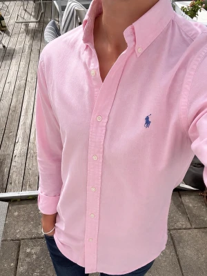 Ralph Lauren Classic Fit Rosa Skjorta  - Stilren Ralph Lauren Classic Fit skjorta i en härlig pastellrosa nyans, med den ikoniska blå hästloggan broderad på bröstet. En tidlös preppy-look med riktig old money-känsla. Tillverkad i ett tunt och luftigt material som passar perfekt året runt. Bär den öppen över en vit T-shirt för en avslappnad look eller knäppt för en mer klassisk stil. Mycket fint begagnat skick. Storlek M.  #RalphLauren #Skjorta #OldMoney #Preppy #Classic