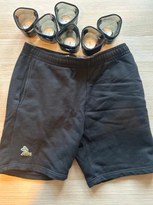 Svarta mjukisshorts med brodyr - Svarta shorts i mjukt material med resår i midjan och sidofickor. Liten broderad detalj i grönt och gult på vänster ben. Perfekta för chill dagar eller träning.