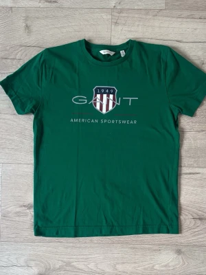 Grön t-shirt från GANT med tryck - Snygg grön t-shirt från GANT med stort tryck framtill och texten 'American Sportswear'. Klassisk rund hals och korta ärmar. Perfekt för dig som gillar sportig och stilren design. Tillverkad i mjuk bomull för skön känsla.