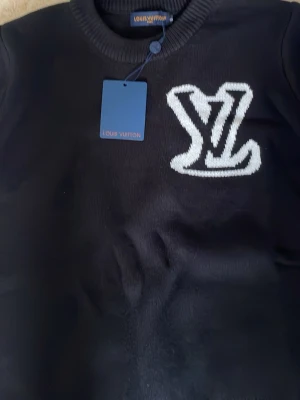 Svart tröja med LV-logga - Snygg svart tröja från Louis Vuitton med stor vit LV-logga på bröstet. Tröjan har rund halsringning och långa ärmar. Perfekt för dig som gillar exklusiva streetwear-plagg och vill sticka ut med en ikonisk design.