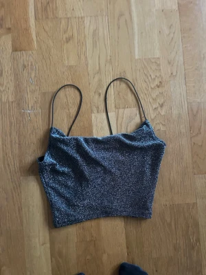 Glittrig svart croppad linnetopp - Snygg glittrig svart linnetopp med tunna axelband och croppad passform. Perfekt för att lyfta vilken outfit som helst med sitt skimrande material och minimalistiska design.