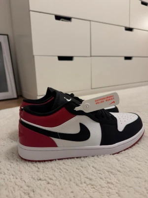 Air Jordan 1 low  - Snygga Nike sneakers i lågt modell med svart, vitt och rött läder. Klassisk swoosh på sidan och perforerad tåbox för extra stil. Skorna har svart snörning och vit mellansula med röd yttersula. Perfekta för dig som gillar streetwear och vill sticka ut. Aldrig använda. 