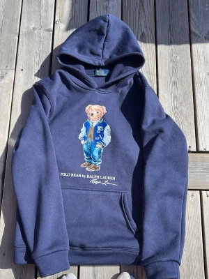 Marinblå hoodie Polo Bear Ralph Lauren - Snygg marinblå hoodie från Ralph Lauren med Polo Bear-tryck på bröstet. Klassisk huva och känguruficka framtill. Mjuk bomullsblandning och avslappnad passform. Perfekt för dig som gillar streetwear och ikoniska prints.