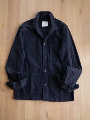 Black A Day’s March Chore Jacket, size L - Stilren svart overshirt i mjuk bomull med klassisk krage och tre fickor framtill. Knäpps med fyra knappar och har en avslappnad passform. Perfekt lager på lager-plagg för en cool och enkel look.