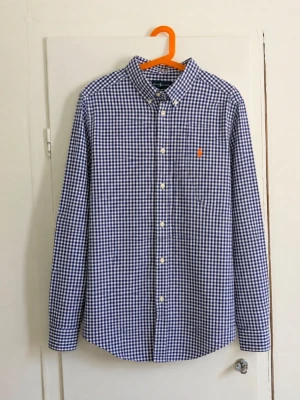 Polo Ralph Lauren rutig skjorta - Långärmad skjorta från Polo Ralph Lauren i blått och vitt med klassiskt små-rutigt mönster. Orange broderad Polo-logga på bröstet och button-down-krage. En stilren skjorta som är enkel att matcha och passar både till vardag och mer uppklätt. 📏 Storlek: L (14-16) ✨ Skick: Bra men den har några fläckar! Hör av dig vid frågor eller om du vill ha mått/fler bilder! 📦