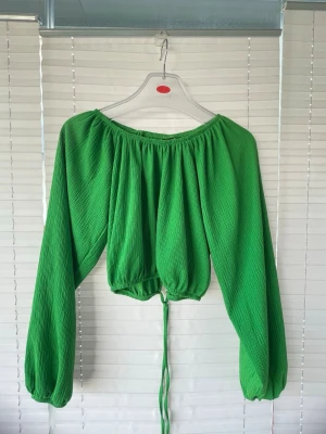 Grön offshoulder blus med knyt - Ryggen är öppen! Trendig grön blus med offshoulder-design och långa, vida ärmar. Toppen har en croppad passform med knytband i midjan för justerbar look. Materialet är lätt och har en strukturerad yta som ger extra stilpoäng.