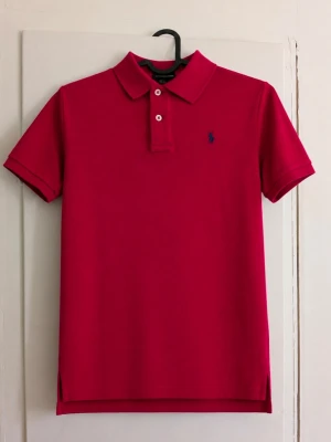 Polo Ralph Lauren röd piké ❤️ - Klassisk röd pikétröja från Polo Ralph Lauren med marinblå broderad logga på bröstet. Kortärmad modell med krage och två knappar framtill. Snygg och tidlös, passar perfekt till jeans, chinos eller shorts. Storlek: M (12-14) Skick: Bra, men den är lite missfärgad i nacken! Skriv gärna vid frågor eller för fler bilder! Pris kan diskuteras vid snabb affär.