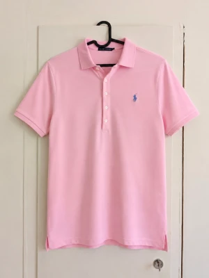 Polo Ralph Lauren rosa piké 🩷 - Ljusrosa pikétröja från Ralph Lauren med klassisk blå broderad logga på bröstet. Kortärmad modell med krage, fyra knappar framtill och små slitsar längst ner. Stilren färg som är perfekt till jeans, shorts eller chinos. Storlek: L Skick: Mycket fint skick utan defekter! Skriv gärna för mått eller fler bilder!