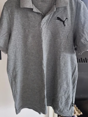 Grå pikétröja från Puma - Klassisk grå pikétröja från Puma med svart logga broderad på bröstet. Tröjan har krage, knappar framtill och korta ärmar. Perfekt till en avslappnad och sportig stil.