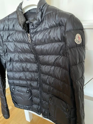 Moncler dunjacka - Snygg mörkblå dunjacka från Moncler med quiltad design och två framfickor med knappar. Jackan har hög krage, dragkedja framtill och Moncler-logga på ärmen. Perfekt för kalla vinterdagar och riktigt lätt att bära. Knappt använt och har kvitto om det behövs. Orginal pris 5495. Perfekt för en kvinna iför hösten