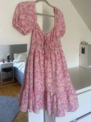 Blommig rosa klänning med puffärm - Söt blommig klänning i rosa och vit med puffärmar och v-ringning. Klänningen har snörning i midjan och volang nedtill. Perfekt för sommarens alla tillfällen och har en luftig, flowy passform.
