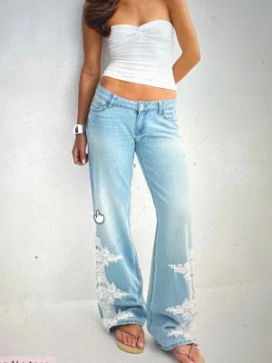 Ljusblå utsvängda jeans med spetsdetaljer - Säljer ett par ljusblå jeans med utsvängda ben och snygga vita spetsapplikationer längst ner på båda benen. Jeansen har klassisk femficksmodell och normal midja. Perfekta för dig som vill sticka ut med en unik och trendig look. Helt nya!