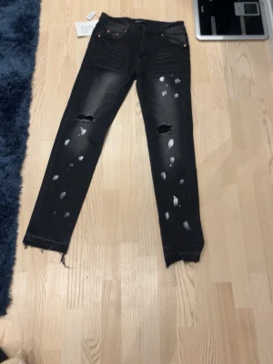 Svarta slitna  jeans - Säljer ett par svarta skinny jeans med slitningar och färgstänk-detaljer. Jeansen har råa, fransiga benslut och hål på knäna för en edgy look. Passar perfekt för dig som gillar streetstyle och vill sticka ut lite extra.