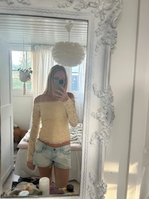 Beige offshoulder spets topp - Säljer en beige offshoulder topp med lång ärm och vacker spetsdetalj. Toppen har en figurnära passform och fina broderade mönster över hela tyget. Perfekt för dig som vill ha en romantisk och trendig look.