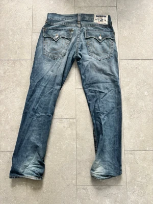 Blå raka true religion jeans - Säljer ett par blå jeans med raka ben och snygga slitningar. Jeansen har klassisk femficksdesign och detaljerade bakfickor med lock och knapp. Materialet är denim i bomull och färgen är tvättad blå med ljusare partier.