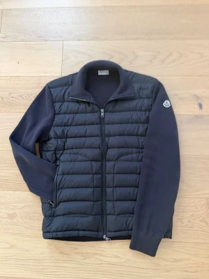 Moncler cardigan - Snygg svart cardiganjacka med quiltad front och stickade ärmar. Jackan har hel dragkedja, ståkrage och Moncler-logga på ena ärmen. Perfekt för lager-på-lager och har två fickor med dragkedja. Materialmix av vadderad polyester och mjuk bomull.