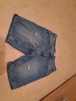 Blå denimshorts med fem fickor - Säljer ett par klassiska blå jeansshorts från LST med fem fickor och knappgylf. De har en loose passform och är tillverkade i slitstark bomull. Perfekta för sommardagar och har en avslappnad look med lite längre ben. I bra skick. Säljs för 40 kr. Tveka inte att kontakta om du är intresserad eller har några frågor! Först till kvarn dom gäller!