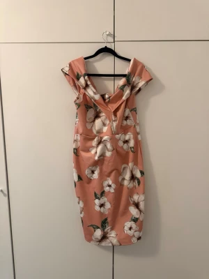Blommig offshoulder klänning - Snygg ljusrosa offshoulder klänning med stort blommönster i vitt och grönt. Klänningen har en figurnära passform och v-ringad urringning med volangdetalj. Perfekt för sommarens alla tillfällen.