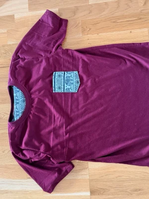 Vinröd t-shirt med mönstrad ficka medium - Säljer en vinröd t-shirt med korta ärmar och en snygg mönstrad ficka på bröstet. T-shirten har rund halsringning med matchande mönster på insidan. Perfekt för dig som vill ha något unikt i garderoben.