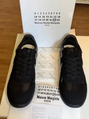 Maison Margiela svarta sneakers - Maison Margiela sneakers i helsvart med klassisk låg silhuett. Skorna har ovandel i läder och mocka, svarta skosnören och diskret logga på hälen. Snygg minimalistisk design som passar till det mesta.