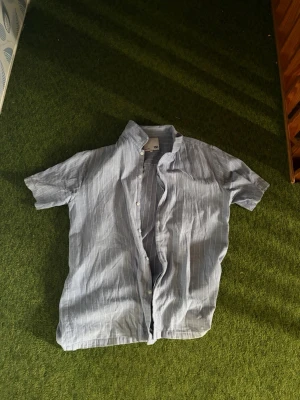 Blåvit randig skjorta från 157 - Kortärmad skjorta från 157 i ljusblått med tunna vita ränder. Skjortan har klassisk krage och knäppning framtill. Perfekt för en avslappnad och stilren look under varmare dagar.
