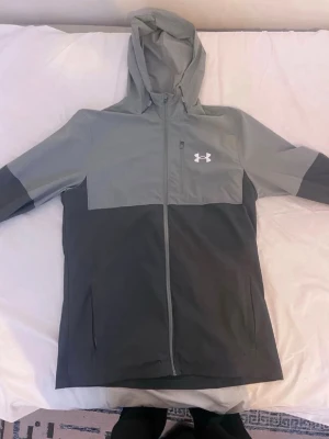 Grå vindjacka från Under Armour - Snygg grå vindjacka från Under Armour med huva och hel dragkedja framtill. Jackan har logga på bröstet och dunder fickor på sidorna. Tvär bra kvalitet, skickar fort och pris går att ta ett snack om!