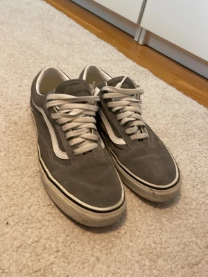 Gråa låga sneakers med vit sula - Snygga grå sneakers med vit sula och vita snören. Skorna har en klassisk låg siluett och en vit sidostripes längs sidan. Ovandelen verkar vara i mocka och textil, vilket ger en skön och stilren look.