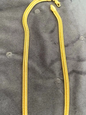 Gult kedjehalsband i metall - Snyggt halsband i guldfärgad metall med platt, tätt sammanlänkad kedja som ger ett exklusivt intryck. Halsbandet har en klassisk karbinhake och passar perfekt för att lyfta vilken outfit som helst. Kedjan har en elegant, glänsande finish och känns rejäl men ändå stilren.