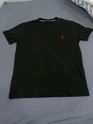 Svart t-shirt från Polo Ralph Lauren - Klassisk svart t-shirt från Polo Ralph Lauren med röd broderad logga på bröstet. T-shirten har rund hals och korta ärmar, perfekt till en enkel och stilren look. Tillverkad i mjuk bomull för skön komfort.