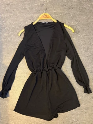Svart långärmad jumpsuit Boohoo - Snygg svart jumpsuit från Boohoo med långa ärmar och djup V-ringning. Modellen har resår i midjan för en skön passform och korta ben. Perfekt för dig som vill ha en enkel men trendig look. Materialet känns mjukt och stretchigt.