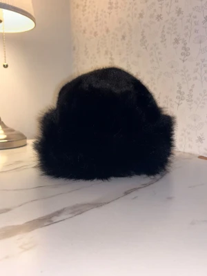 Svart fluffig pälsmössa - Säljer en svart pälsmössa med mjuk och fluffig känsla. Mössan har en klassisk rund form och är helt täckt av glansig fuskpäls som ger en lyxig look. Perfekt för kalla dagar när du vill hålla värmen och ändå se snygg ut. Använd 2 gånger. 