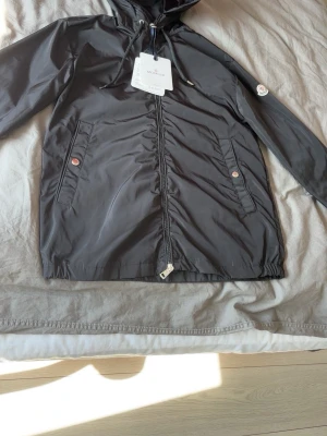 Mon”cler windbreaker  - Snygg svart vindjacka från Mon”cler med huva och dragkedja framtill. Jackan har två fickor med knappstängning och klassisk Moncler-logga på ärmen. Tillverkad i lätt, glansigt material som står emot vind. Perfekt för en trendig och sportig look.