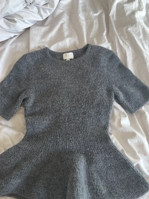 Grå stickad topp med peplum - Snygg grå stickad topp med peplumdetalj och korta ärmar. Rund halsringning och mjukt, ribbat material som ger en skön känsla. Perfekt för dig som vill ha en trendig och bekväm look till vardags.