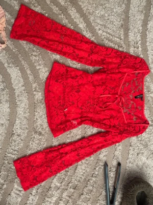 Röd spetsblus med knyt från Pieces - Säljer en röd långärmad spetsblus från Pieces med blommigt mönster och snygg knytning framtill. Blusen har en figurnära passform och transparent spets som ger en trendig look. Perfekt att styla med jeans eller kjol.