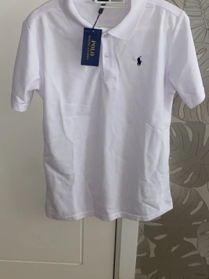 pikétröja från Polo Ralph Lauren - Klassisk vit pikétröja från Polo Ralph Lauren med korta ärmar och blå logga broderad på bröstet. Tröjan har krage och knäppning framtill. Perfekt till jeans eller shorts för en clean och stilren look.