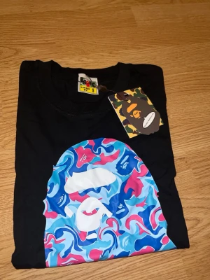 Svart BAPE t-shirt  - Svart t-shirt från BAPE med ikoniskt ape-head tryck i blått, rosa och vitt framtill. Klassisk passform och rund hals. Materialet är mjuk bomull och t-shirten har korta ärmar. Perfekt för dig som gillar streetwear och vill sticka ut.