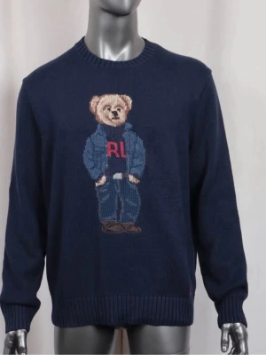 Ralph Lauren tröja - Mörkblå stickad tröja från Ralph Lauren med en stor broderad björn på bröstet. Björnen har på sig jeans, jacka och en tröja med röda bokstäver. Tröjan har rund halsringning och ribbade muddar vid ärmslut och nederkant. Perfekt för dig som gillar unika detaljer.