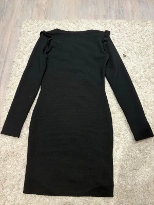 Svart långärmad klänning med volang - En svart, figurnära klänning med långa ärmar och volangdetaljer vid axlarna. Klänningen har en enkel, stilren design och är tillverkad i ett mjukt material som formar sig efter kroppen. Perfekt för dig som gillar minimalistisk stil med en twist.