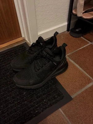 Nike Air Max 270 helsvarta sneakers - Snygga helsvarta Nike Air Max 270 sneakers med mesh-överdel och stor synlig Air-enhet i hälen. Skorna har en sportig siluett, diskret Swoosh-logga och dragflik bak. Perfekta för dig som gillar stilrena och bekväma sneakers med modern känsla.