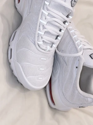 Vita Nike Air Max Plus sneakers - Säljer ett par fräscha vita Nike Air Max Plus sneakers med mesh- och syntetmaterial, ikoniska vågmönster och synlig Air-dämpning i sulan. Skorna har snörning och en sportig, chunky look som sticker ut.