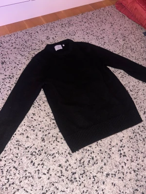Svart stickad tröja från Bläck - En klassisk svart stickad tröja från Bläck i storlek S. Tröjan har rund halsringning, ribbade muddar vid ärmslut och nederkant samt lång ärm. Perfekt för lager-på-lager och enkel att matcha med allt.