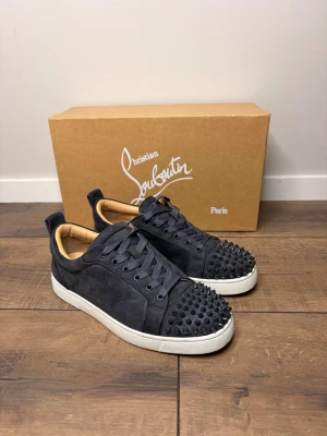 Christian loubutin skor  - Unika svarta sneakers med mattsvarta nitar på tån och ikonisk röd sula. Skorna har svarta skosnören, vit yttersula och är tillverkade i mockaliknande material. Perfekt för dig som vill sticka ut med en edgy och trendig look. Obs endast skorna ingår 