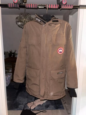 Brun canada goose jacka med fuskpälsluva - Säljer en brun canada goose jacka med luva och fuskpälsdetalj. den har flera praktiska fickor framtill, dragkedja och kardborrestängning. Märke med logga på bröstet och justerbar huva. Perfekt för kalla dagar och riktigt skön att bära.