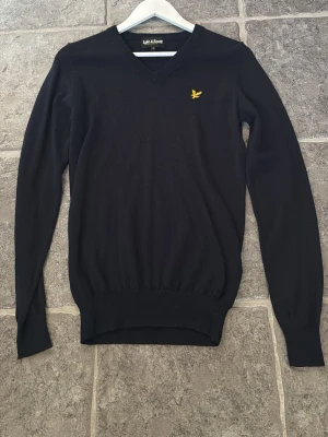 Mörkblå stickad tröja Lyle & Scott - Snygg mörkblå stickad tröja från Lyle & Scott med V-ringning och den klassiska gula logon broderad på bröstet. Tröjan har långa ärmar och ribbade muddar vid ärmslut och nederkant. Perfekt till lager på lager och enkel att matcha.